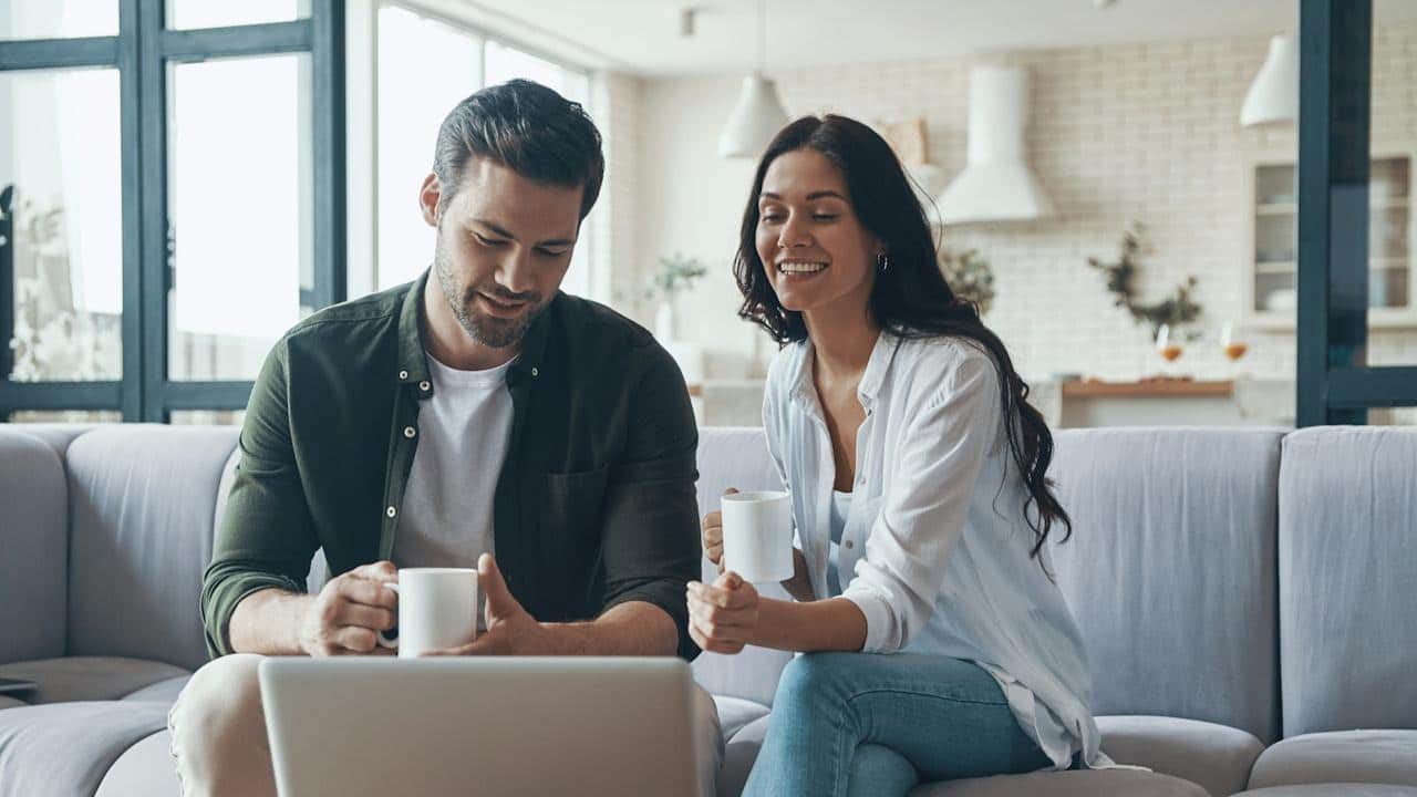 A beautiful young couple drinking coffee and looking at the laptop while sitting on the sofa at home.