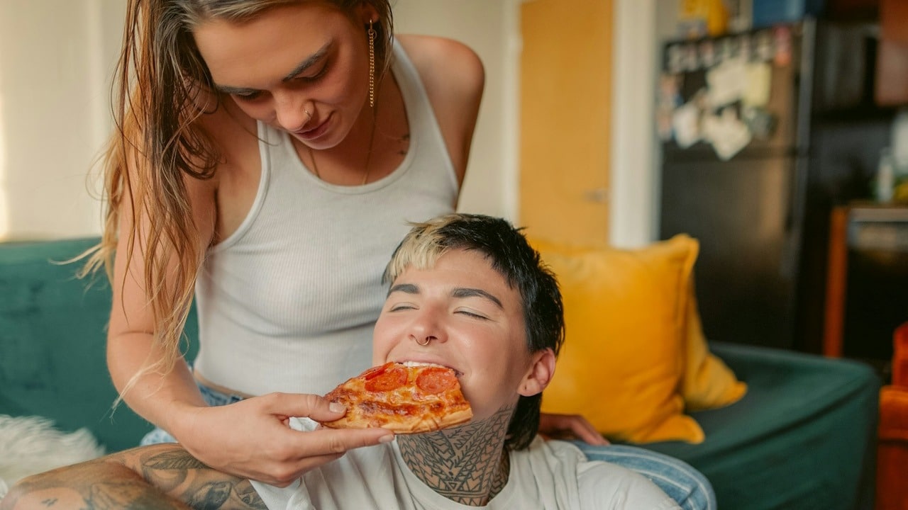 A woman sitting on a couch feeding a slice of pizza to a man who sits while leaning against her legs.