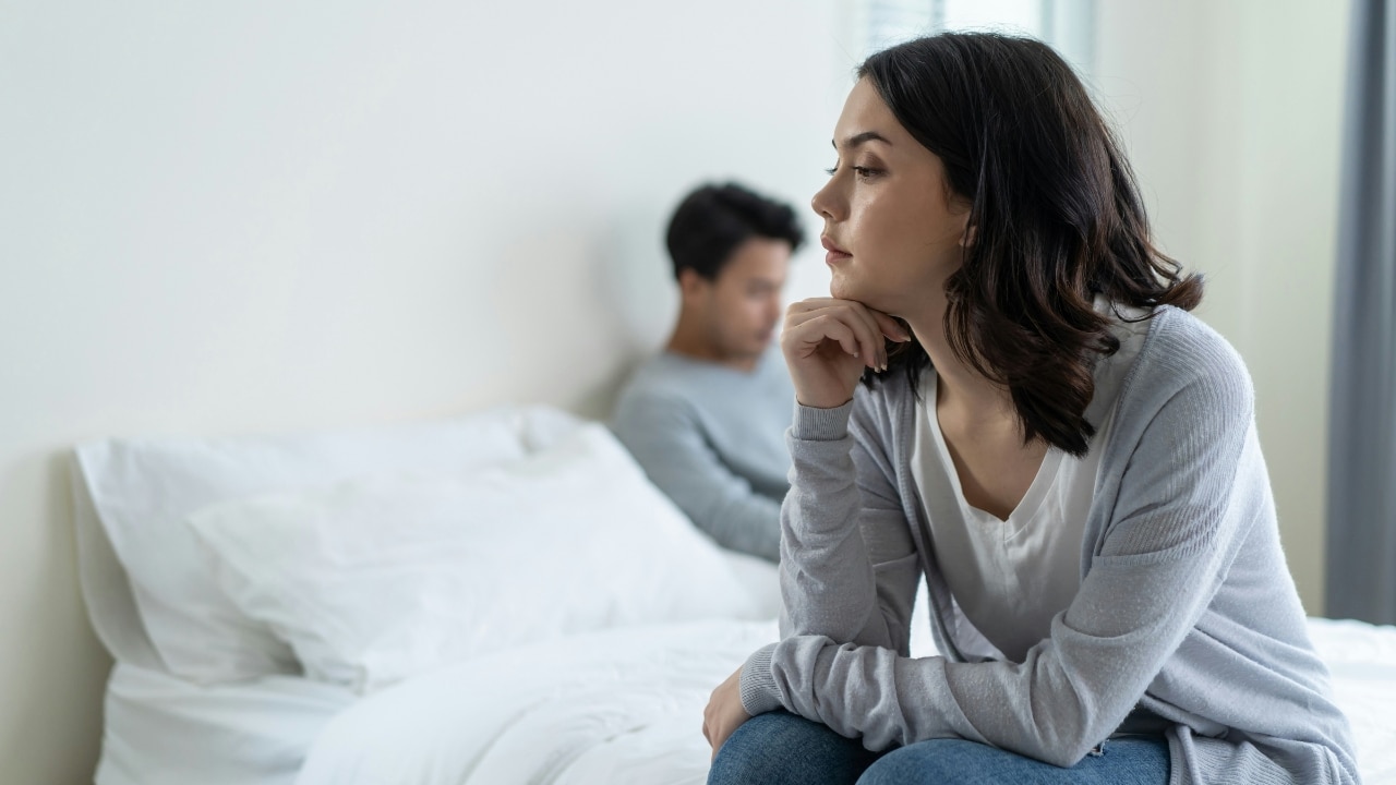 A woman sits on a bed looking away while a man sits blurred in the background.