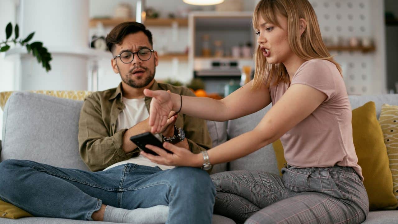 A woman gestures toward a smartphone while sitting on a couch next to a man.