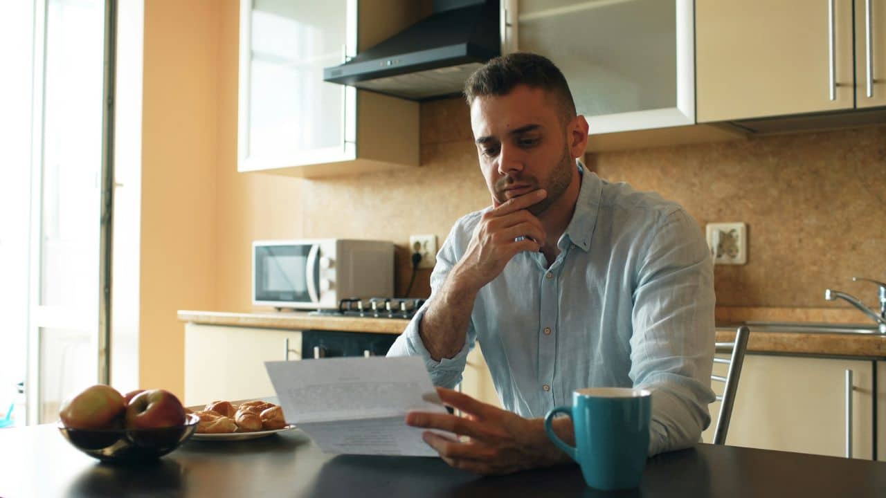 A man sits at a kitchen table holding a piece of paper and looking down.