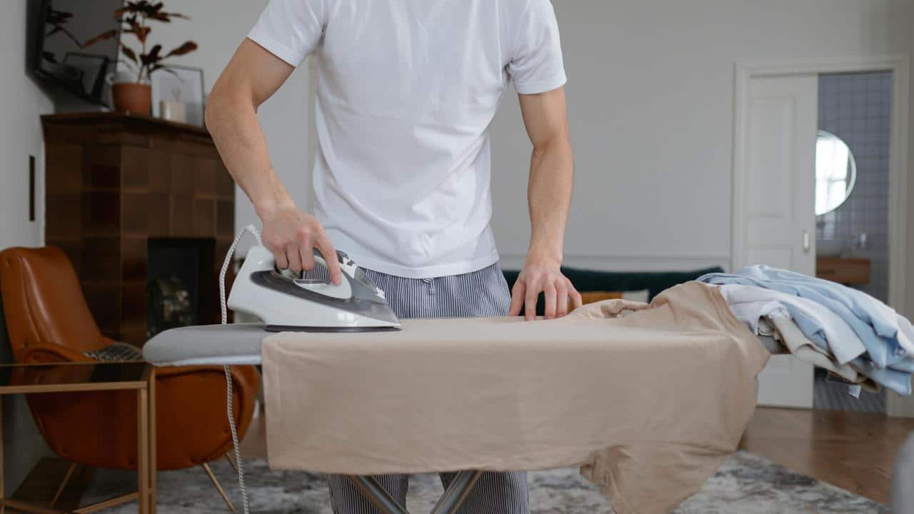 A person ironing a beige shirt on an ironing board in a tidy room.