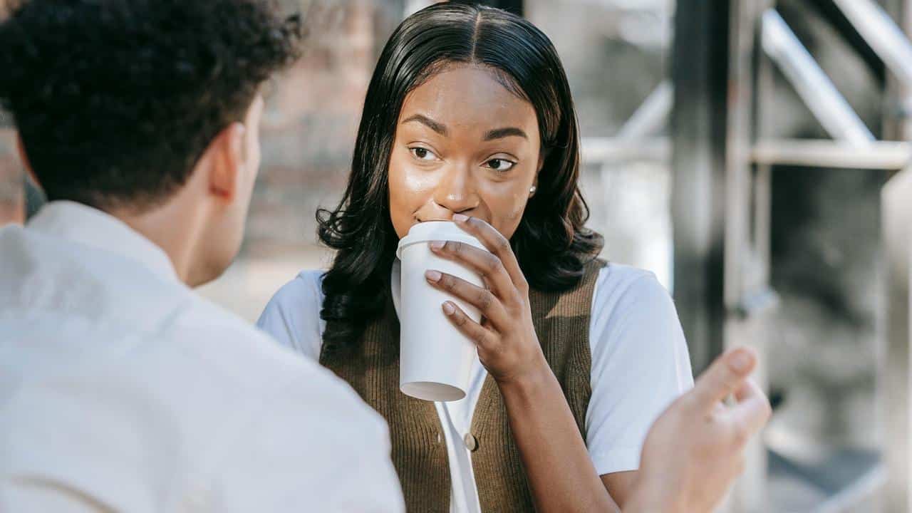 A woman drinking coffee while talking to a man at an outdoor café.