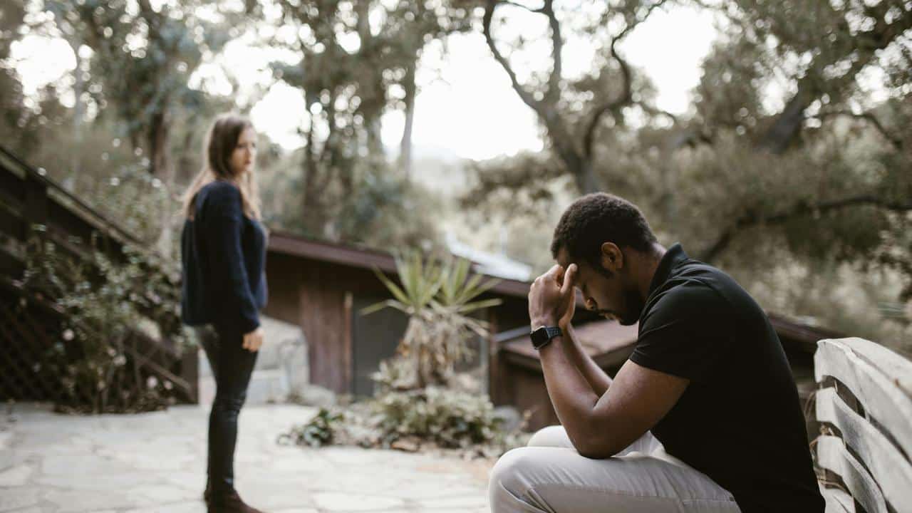 A man sitting with his head in his hands while a woman stands nearby looking upset.
