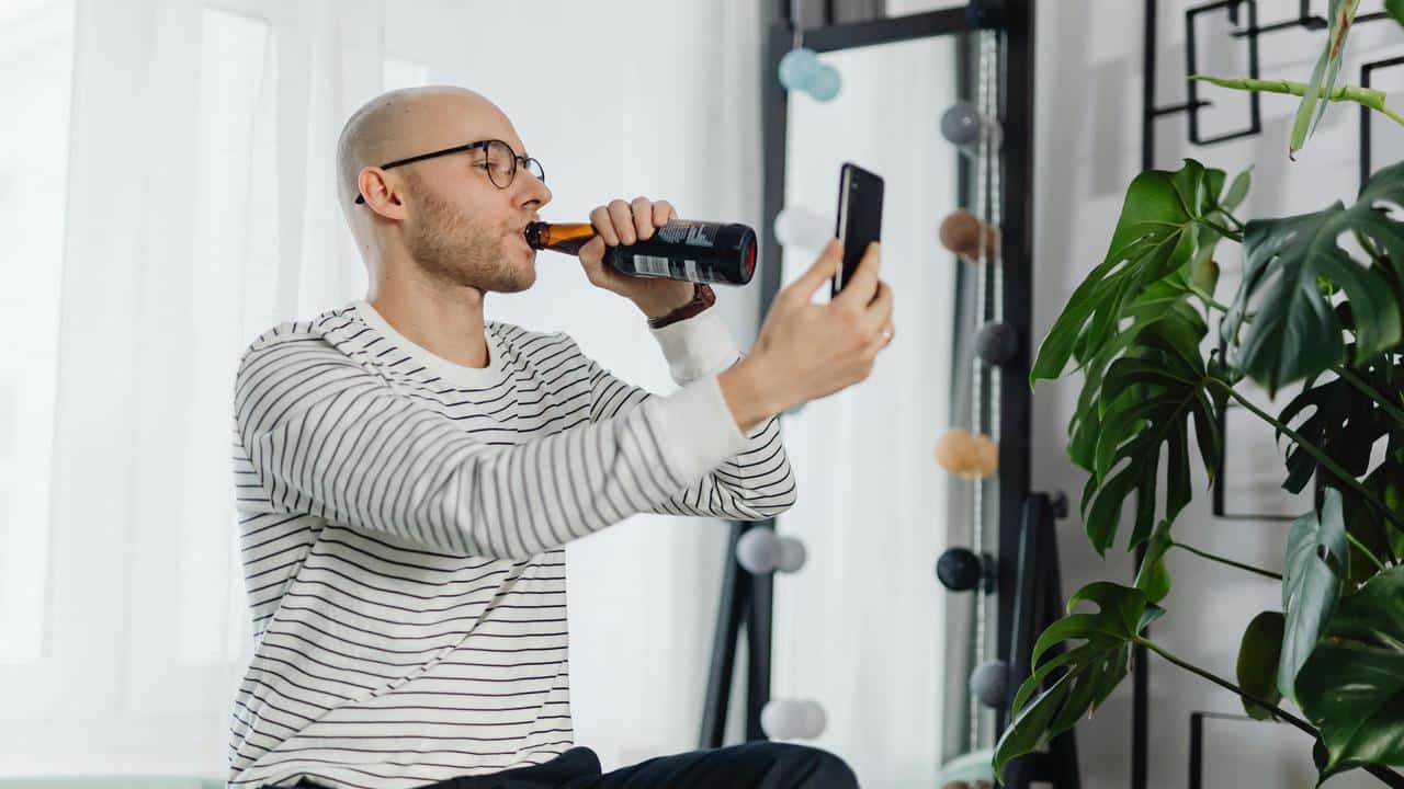 A man drinking from a bottle while holding a phone indoors.