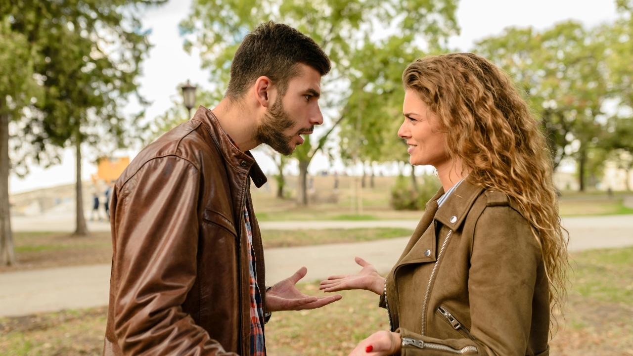 A man in a leather jacket and a woman in a green jacket gesture outdoors.
