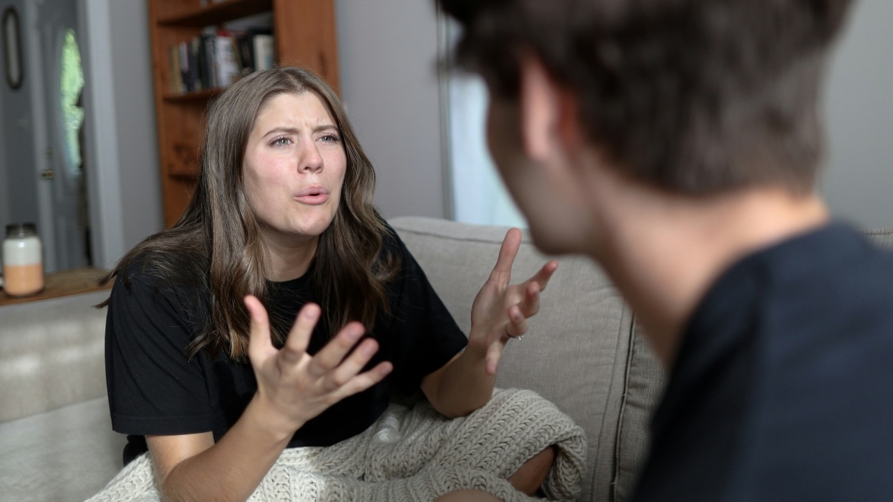 A woman with long hair gestures with both hands while speaking to a man’s blurred profile.