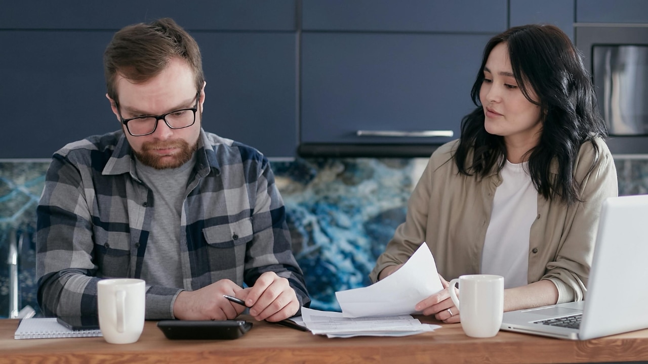 A man and woman sit at a counter reviewing papers with a laptop and mugs.