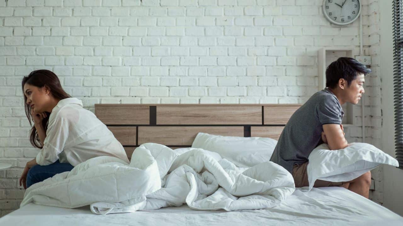A man and woman sit on opposite sides of a bed looking away toward different walls.