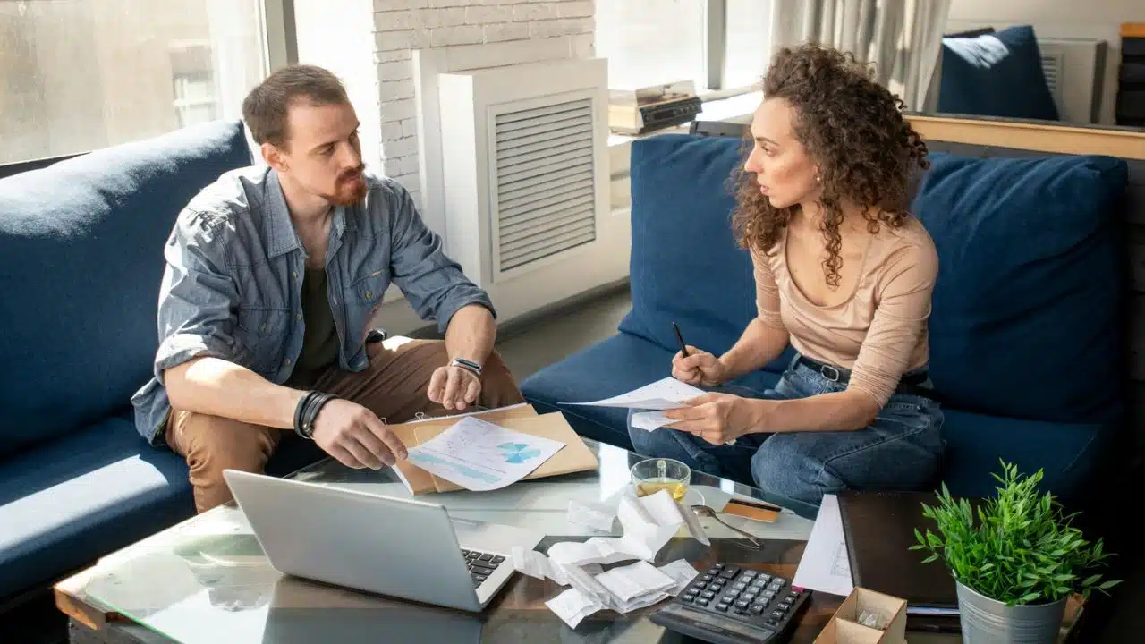 A man and woman sit on a sofa reviewing charts, receipts, and a laptop.