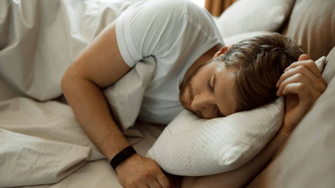 A man with light brown hair and a beard sleeps on a pillow in a white bed.