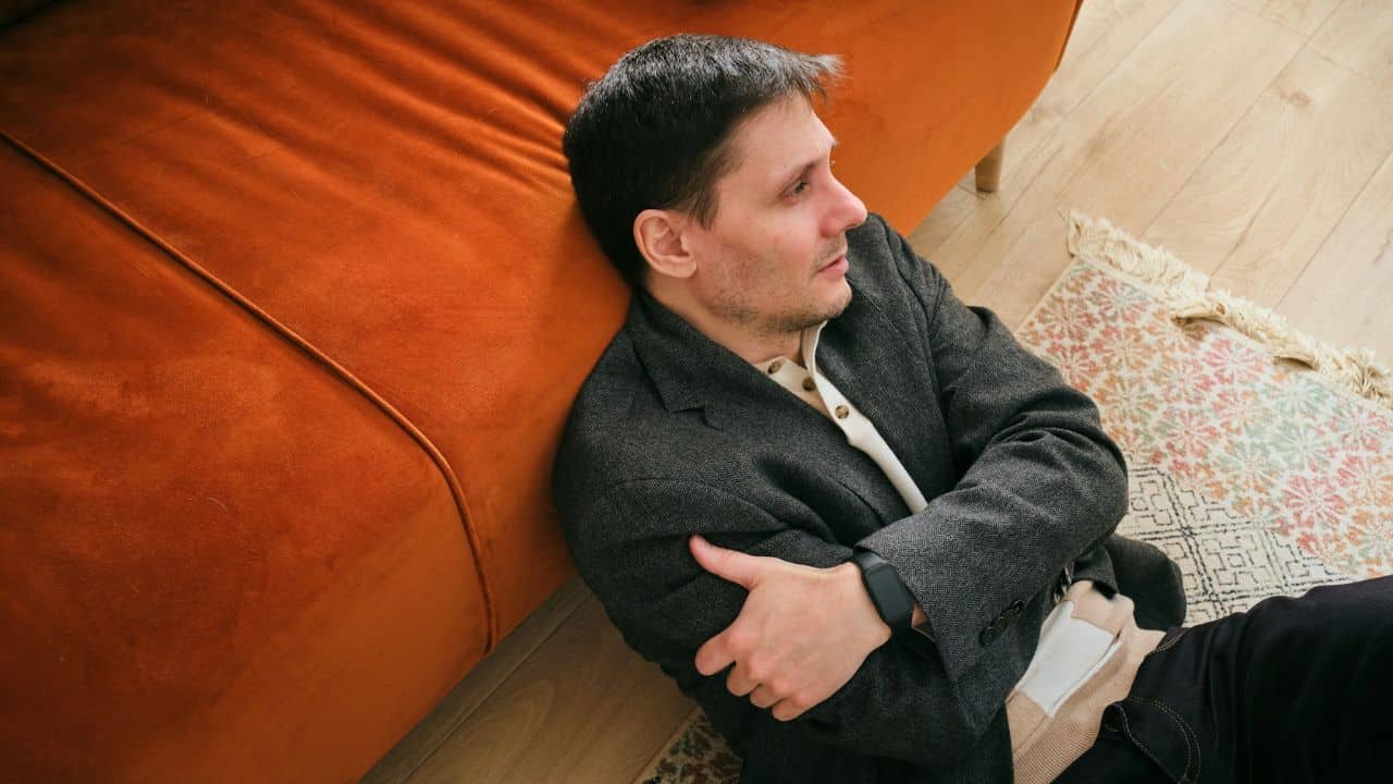 Man in a grey blazer sitting on the floor, leaning against an orange sofa with arms crossed.