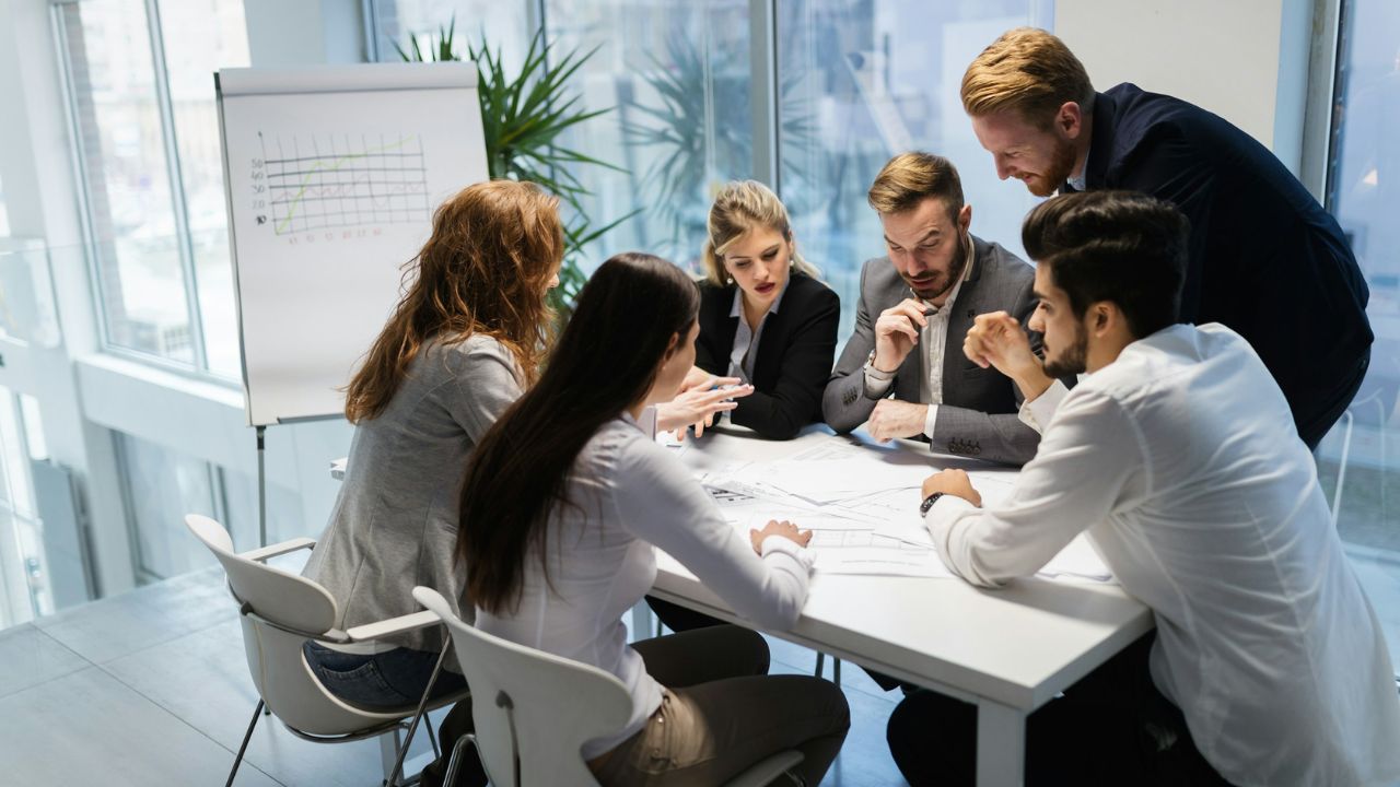 Six professionals gathered around a white conference table in a bright, modern office during a meeting.