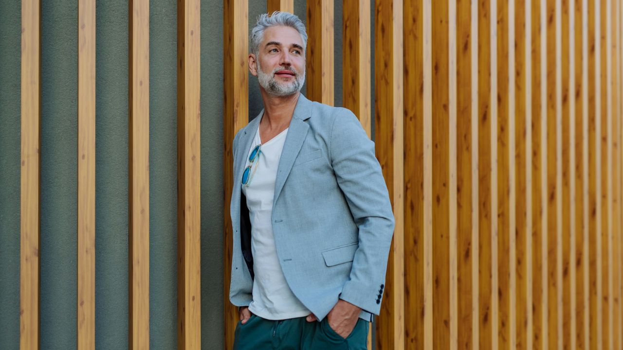 Stylish middle-aged man with gray hair posing against a vertical wood-slat wall.