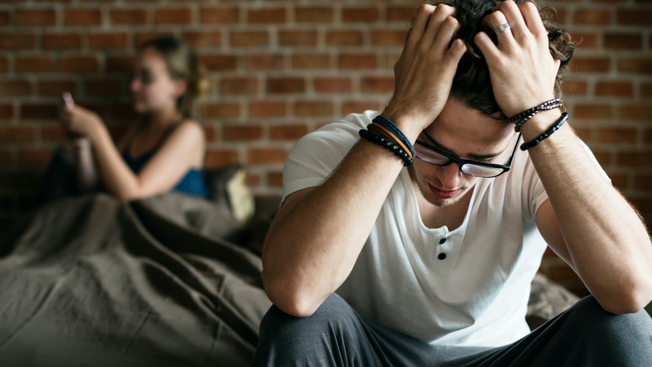 A stressed man wearing glasses sits on a bed, holding his head, while a woman in the background looks at her phone.