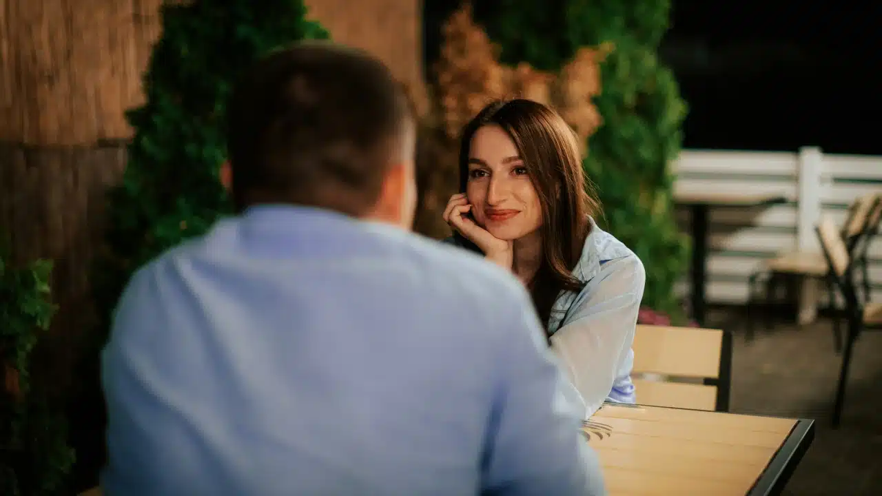 A woman rests her chin on her hand while looking at a man across a table.