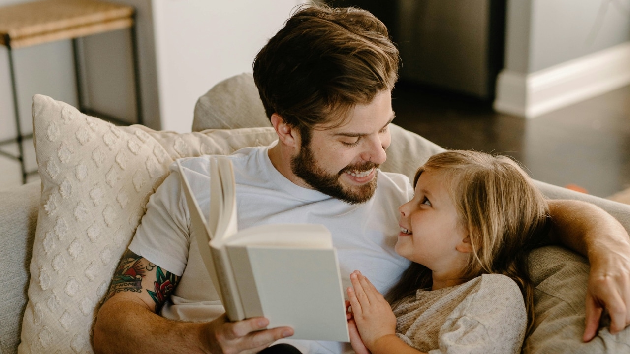 A smiling man with tattoos holds an open book while looking at a young girl.