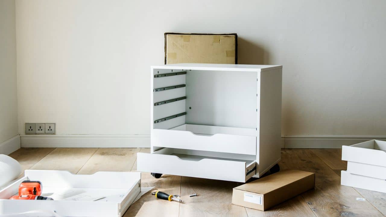 A partially assembled white cabinet sits on a wooden floor surrounded by tools and boxes.