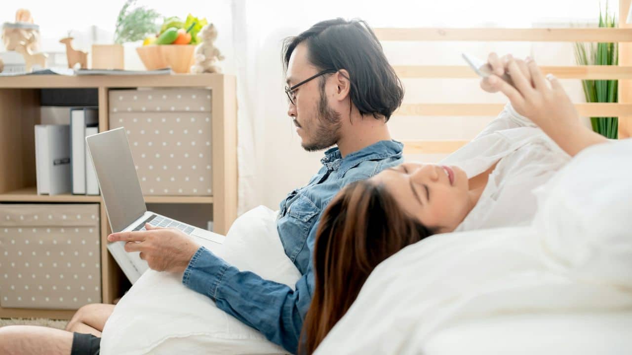 A man uses a laptop while a woman lies beside him looking at a phone.