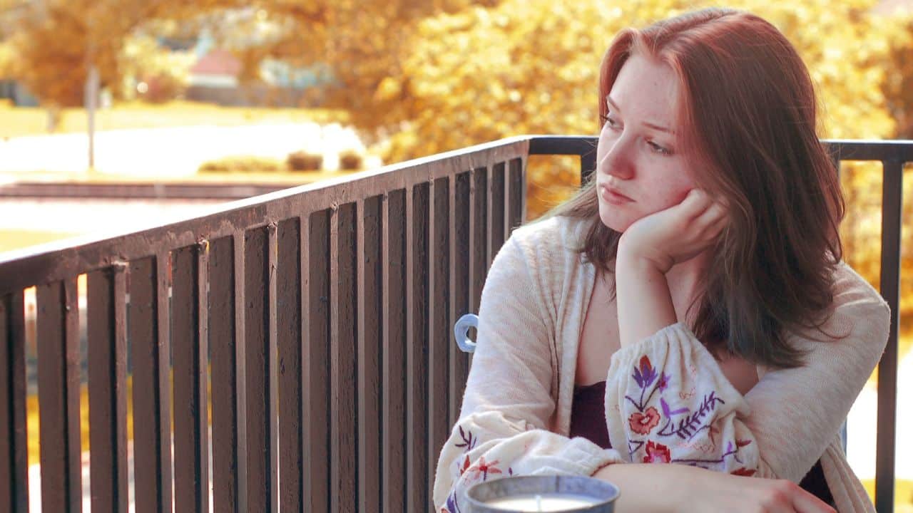 A woman with reddish hair sits on a balcony, resting her chin on her hand.