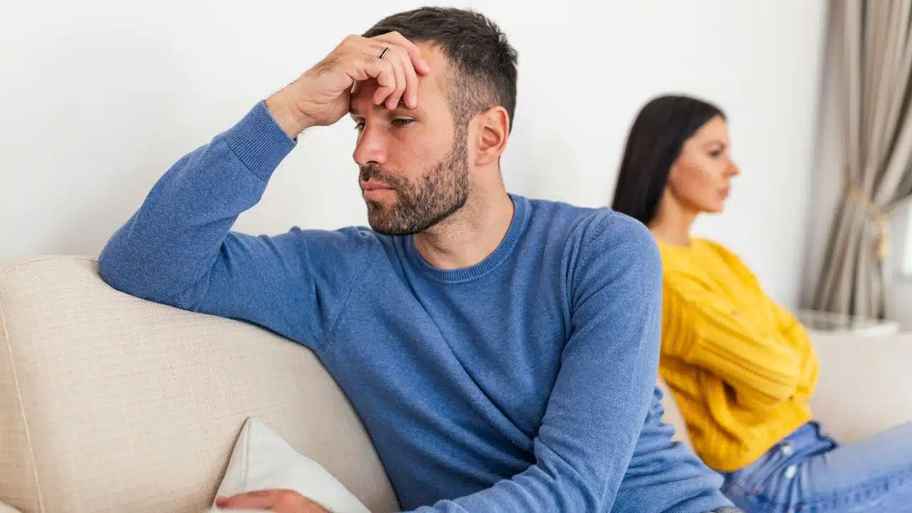 A man in a blue sweater touches his forehead while a woman sits behind him.