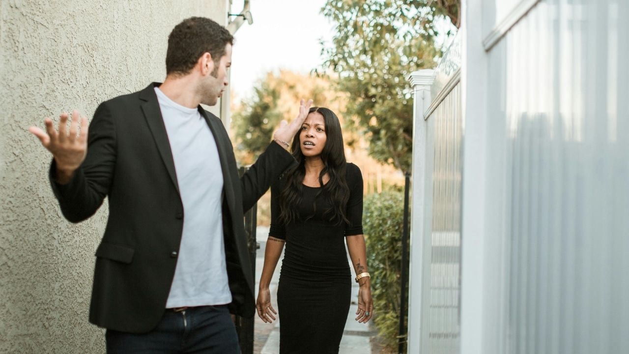 A man and woman stand outdoors near a white fence, appearing to have a tense conversation.