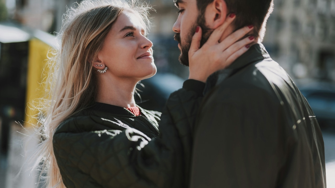 A woman smiles while holding a man's face with both hands in a close-up shot.