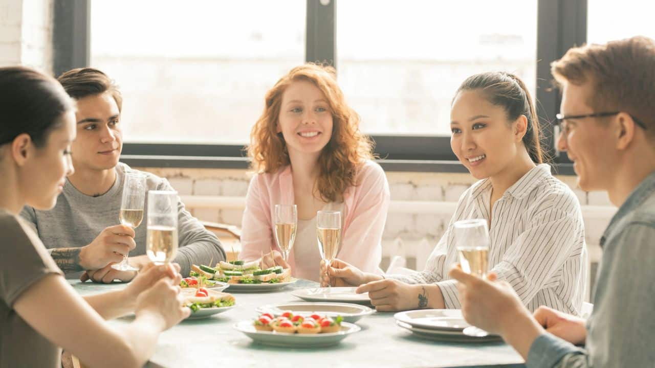 A group of five people sit around a table with snacks and glasses of wine.