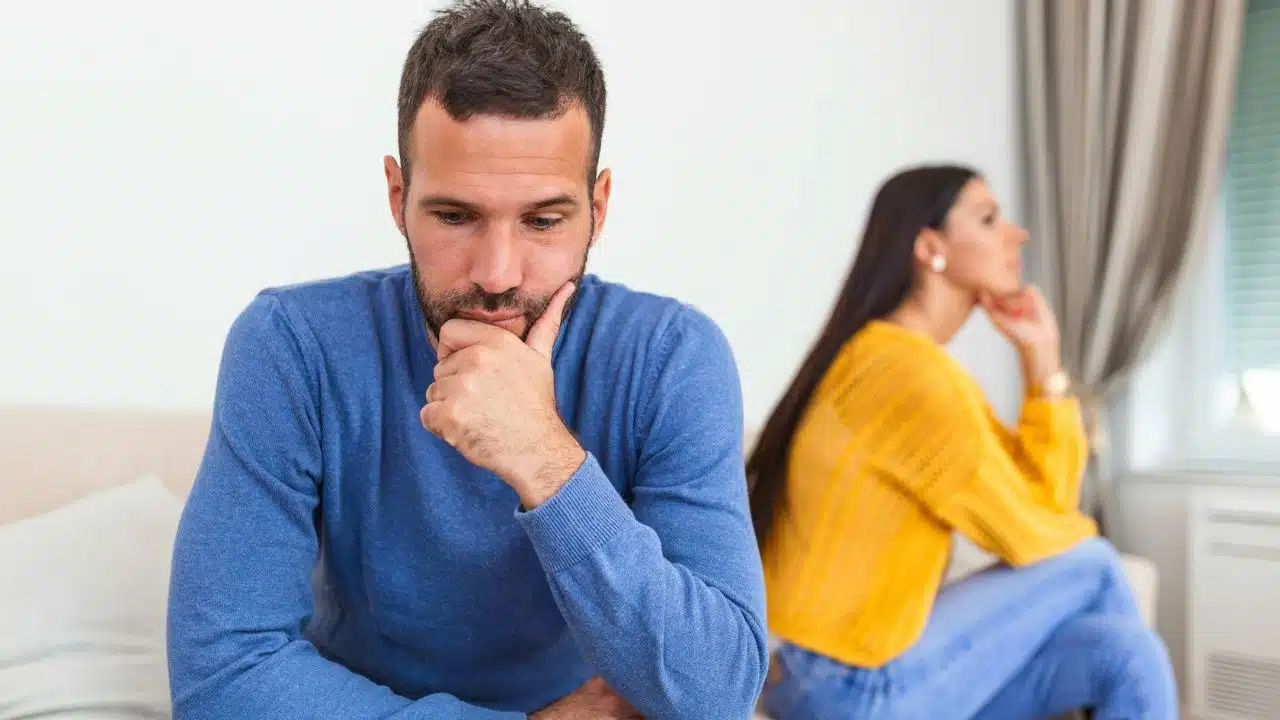 A man in a blue sweater looks down while a woman looks away in the background.