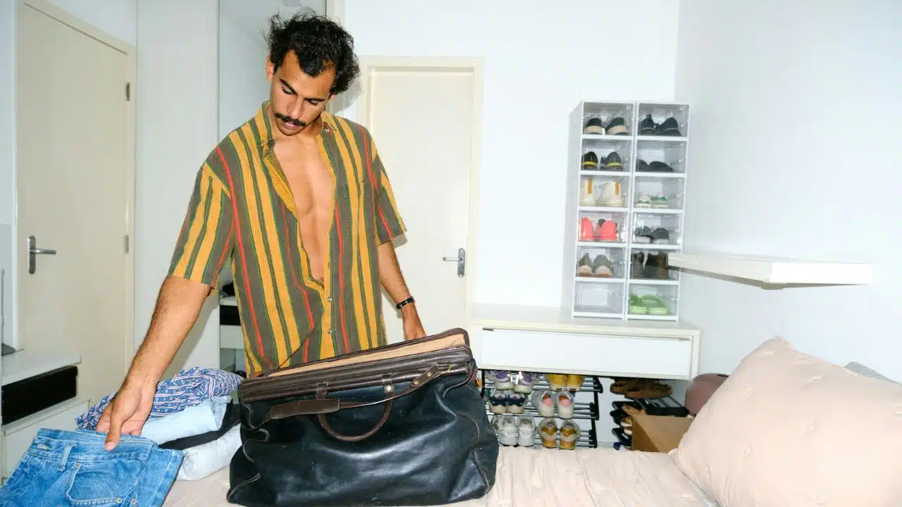 A man in a striped shirt packs clothing into a large black bag on a bed.