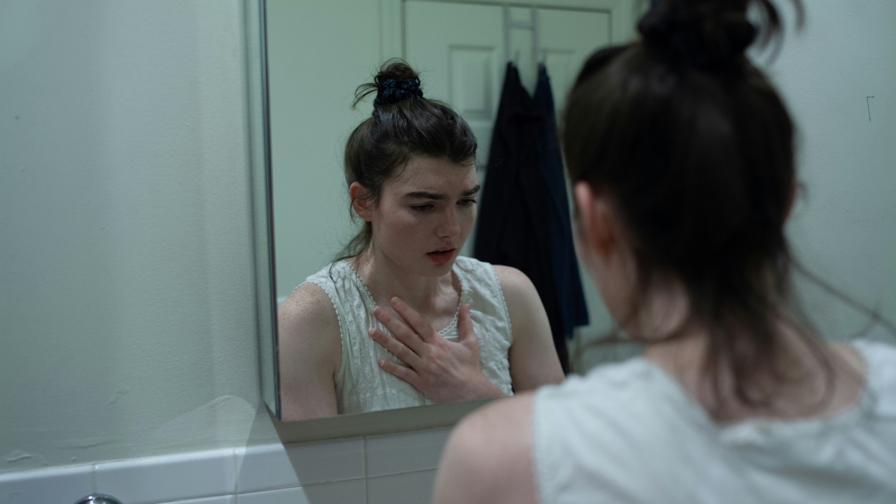 A woman with her hair in a bun looks into a mirror with her hand on her chest.