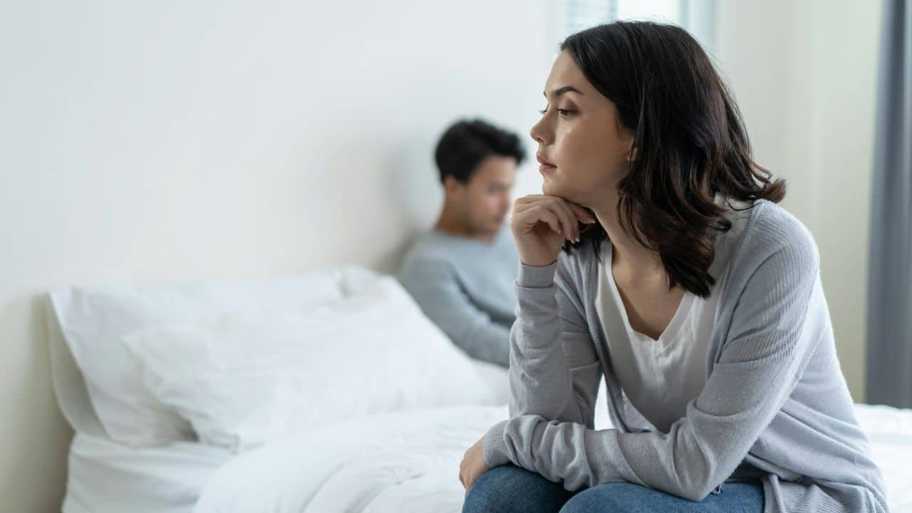 A woman sits on the edge of a bed while a man sits blurred behind her.