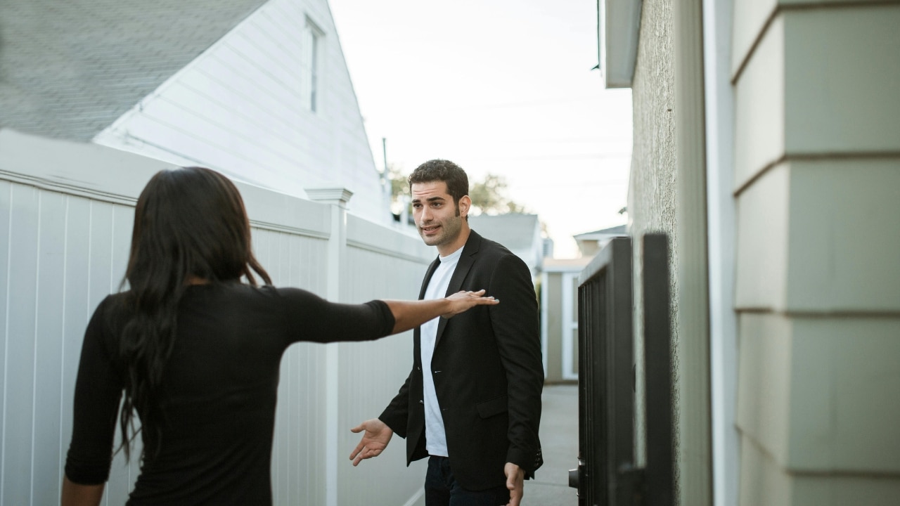 A woman in black gestures toward a man standing in an outdoor walkway between buildings.