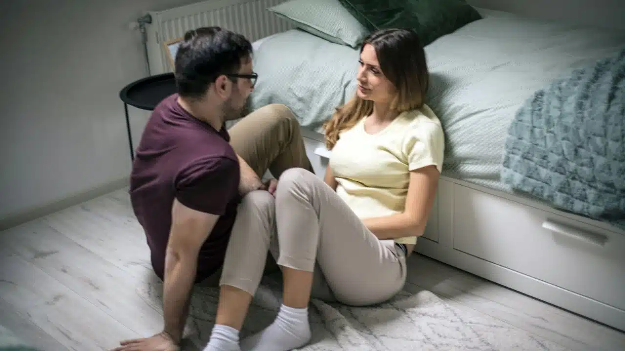 A man and woman sit on a bedroom floor facing each other and talking.