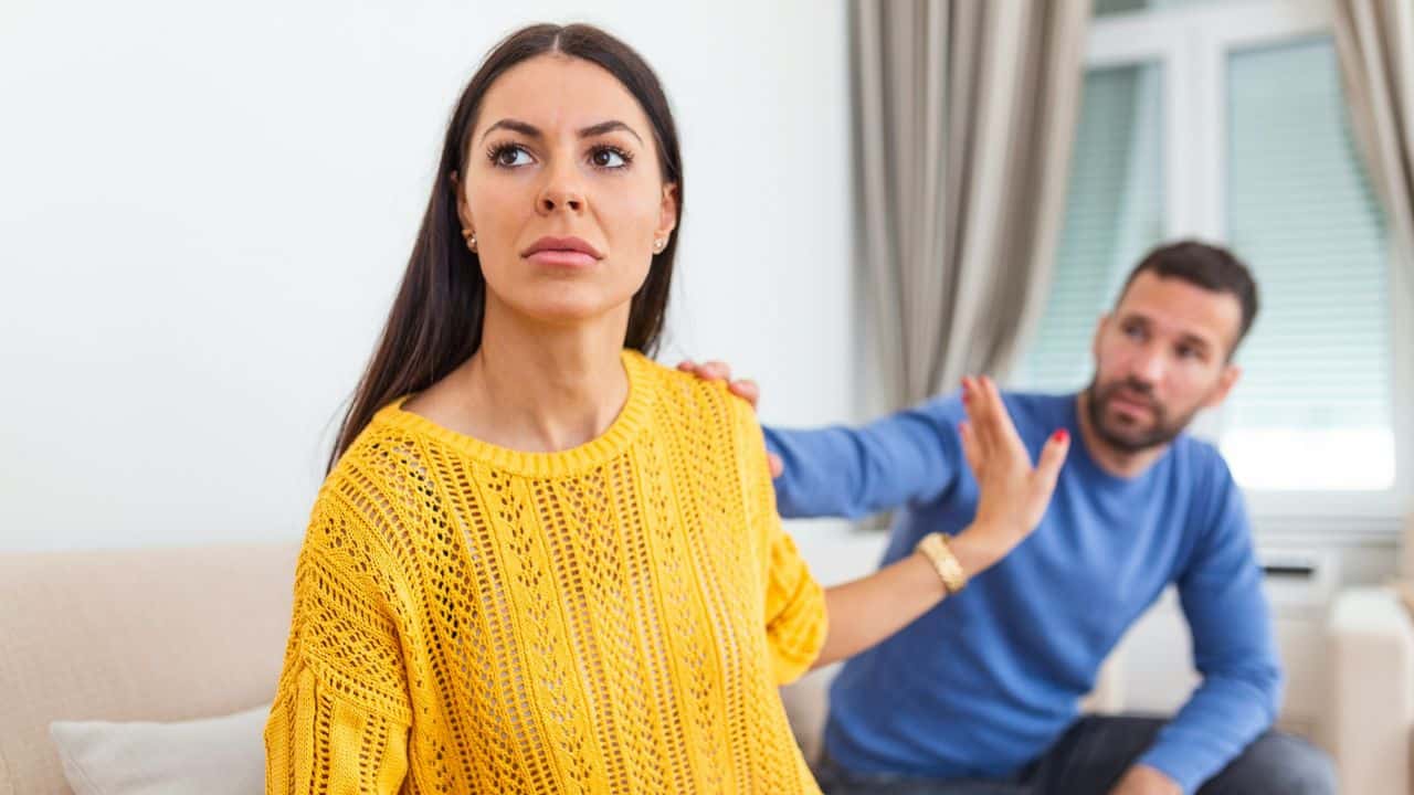 A woman in a yellow sweater looks away while a man behind her reaches out.