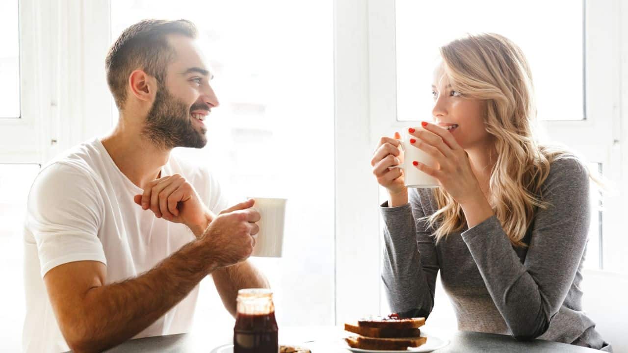 A man and woman sit at a table, smiling and holding white mugs near toast.