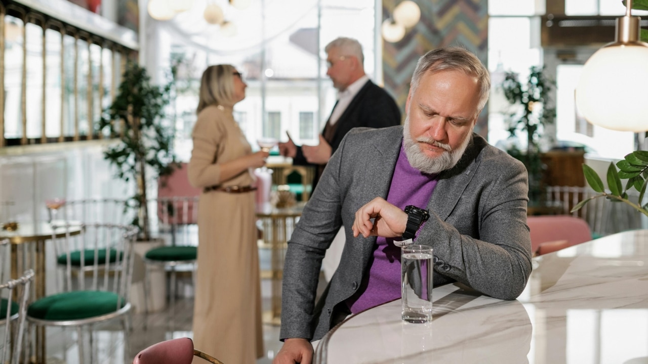 An older man in a grey blazer checks his watch while leaning on a white bar.