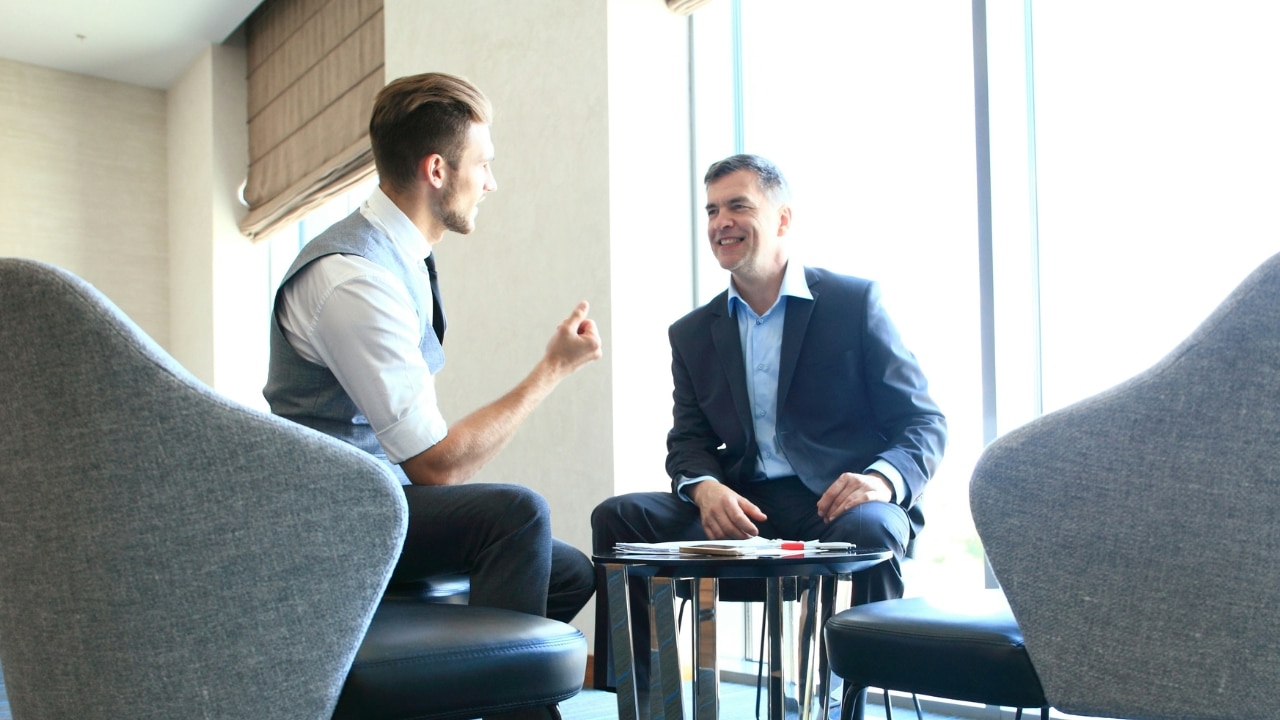Two men in business attire sit in a bright room having a face-to-face conversation.