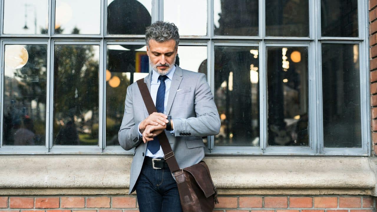 A man in a grey suit and shoulder bag looks down at his wrist watch.
