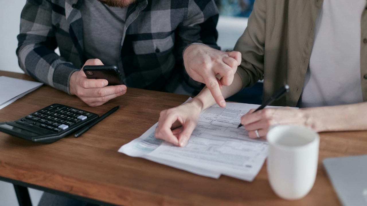 Two people sit at a desk with a calculator, a phone, and a paper document.