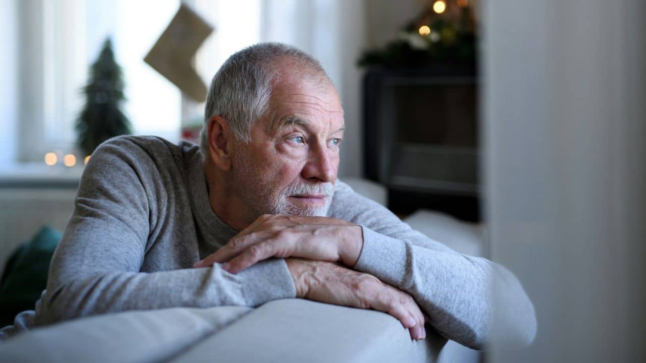 An older man rests his chin on his hands while looking out of a window.