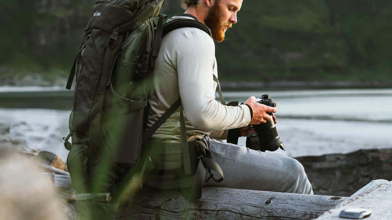 A man with a backpack sitting outdoors holding a camera.