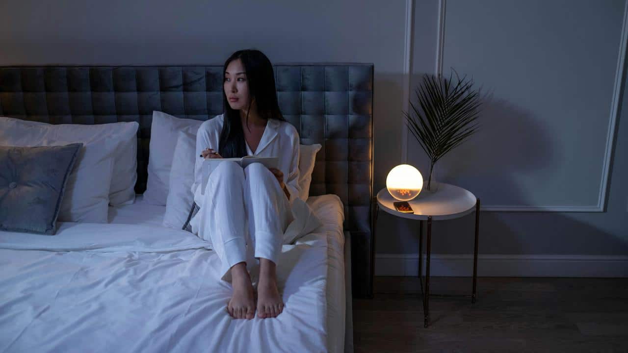 A woman sitting on a bed at night, writing in a notebook.