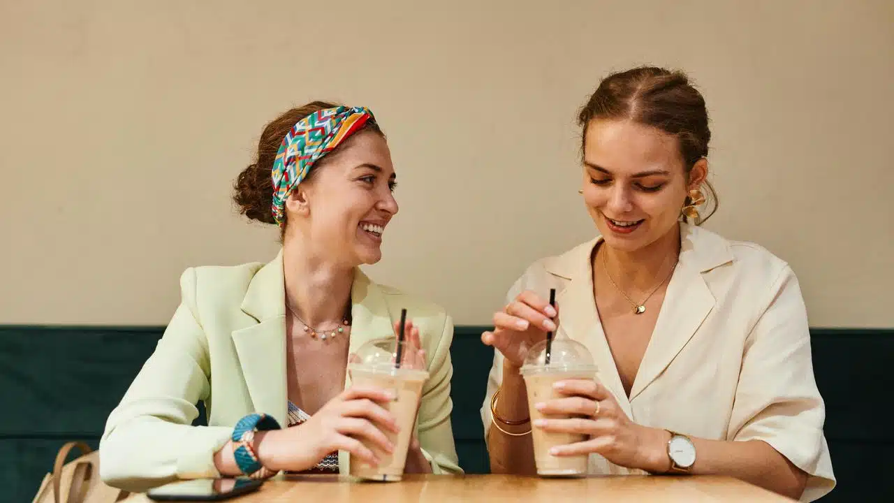 A pair of women smiling and chatting over iced drinks at a café table.