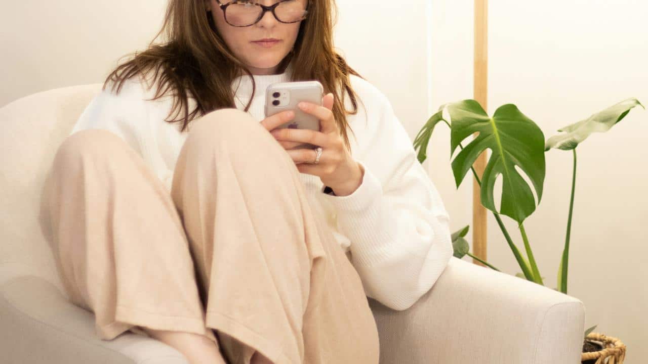 A woman wearing glasses sitting in a chair and looking at her smartphone.