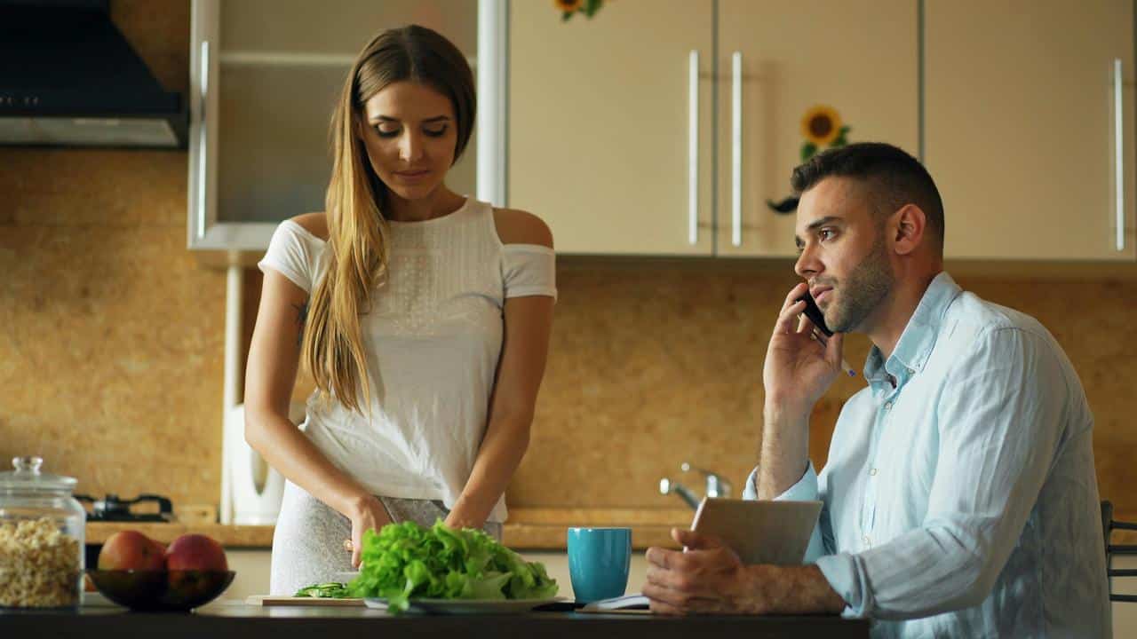 A woman preparing food while a man talks on the phone at a kitchen table.
