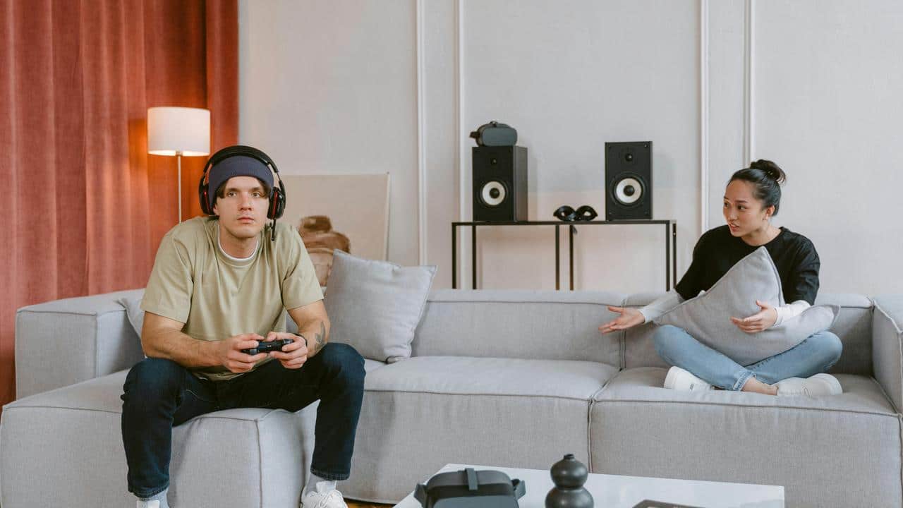 A man gaming while a woman speaks to him on the couch.