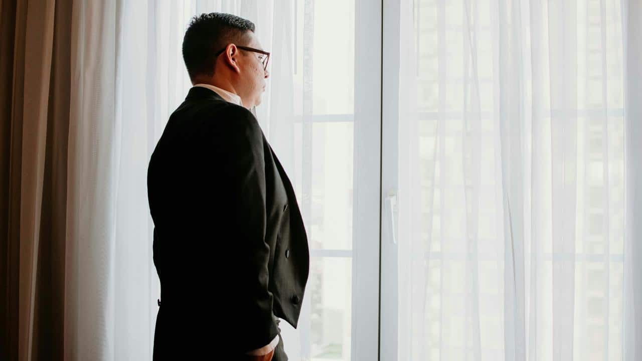 A man in a suit standing by a window and looking outside.