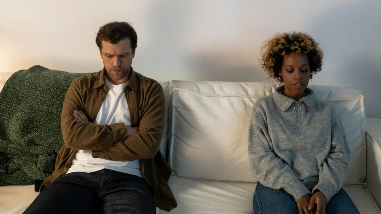 A man and woman sit far apart on a white couch, looking down in silence.