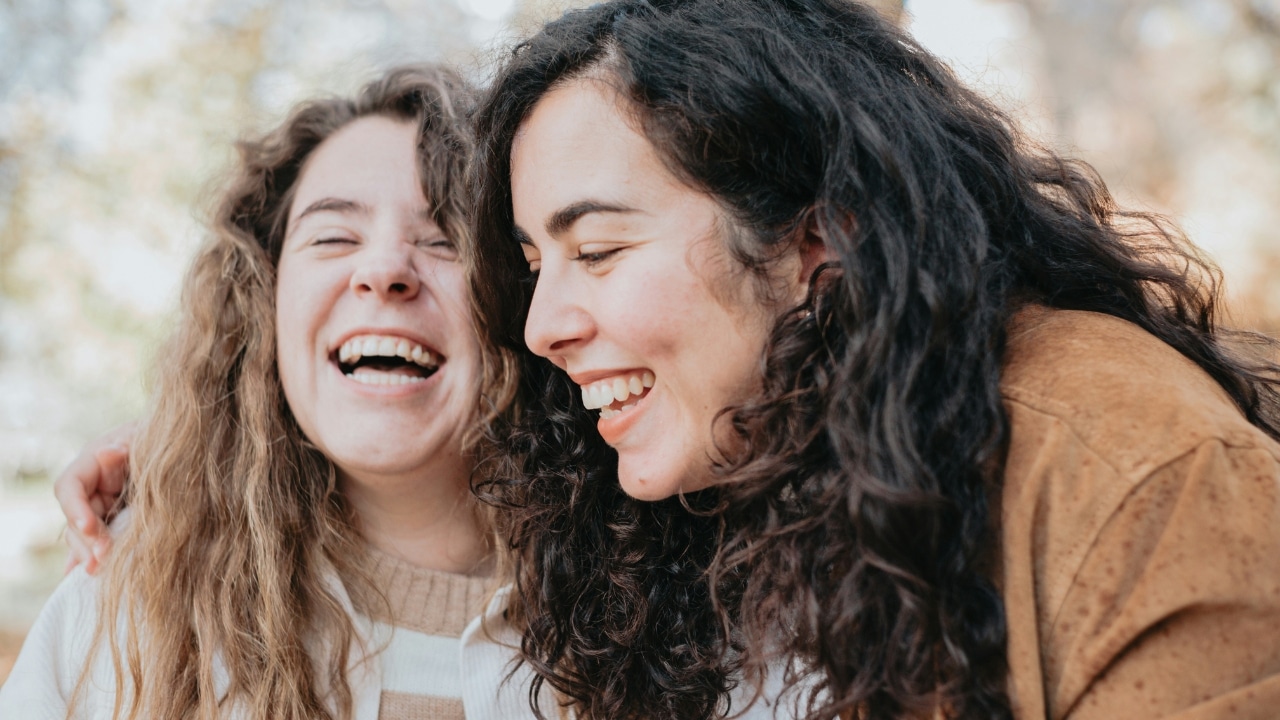 Two women with long curly hair laughing closely together in a bright outdoor setting.